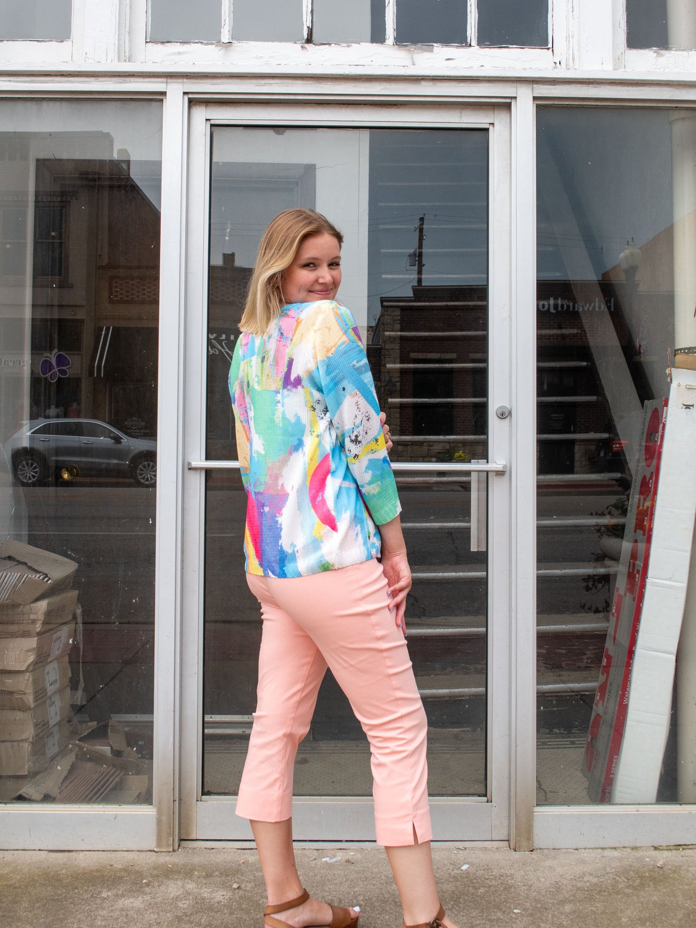 A model wearing a blue colorful patterned, short, knit cardigan. The model has it paired with a white tank, peach pants and brown sandals.
