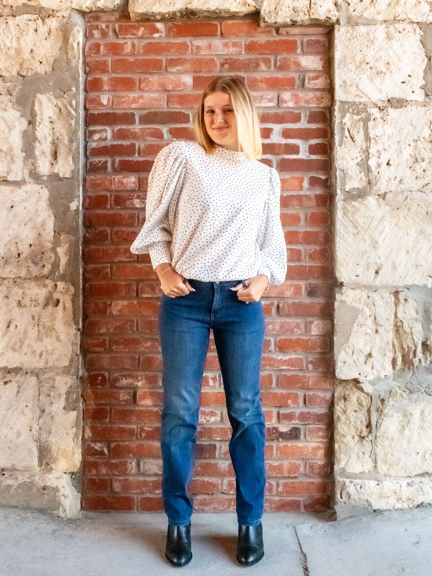 A model wearing a pair of straight leg, dark wash jeans. She has them paired with a white polka dot blouse and black booties.