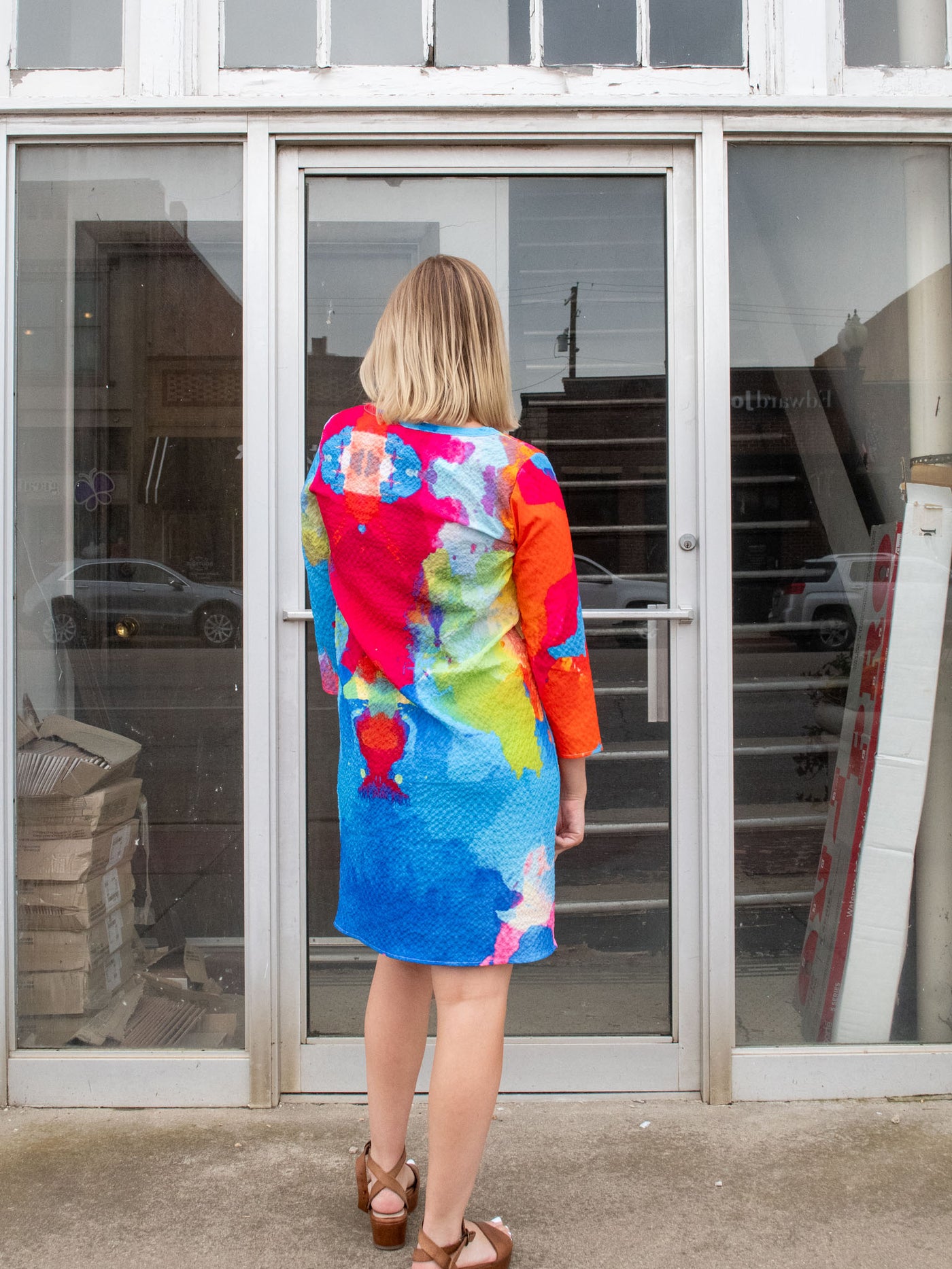 A model wearing a mid thigh length dress that features pink, blue, green, orange, and yellow watercolor details and 3/4 sleeve length. The model paired it with brown platform sandals.