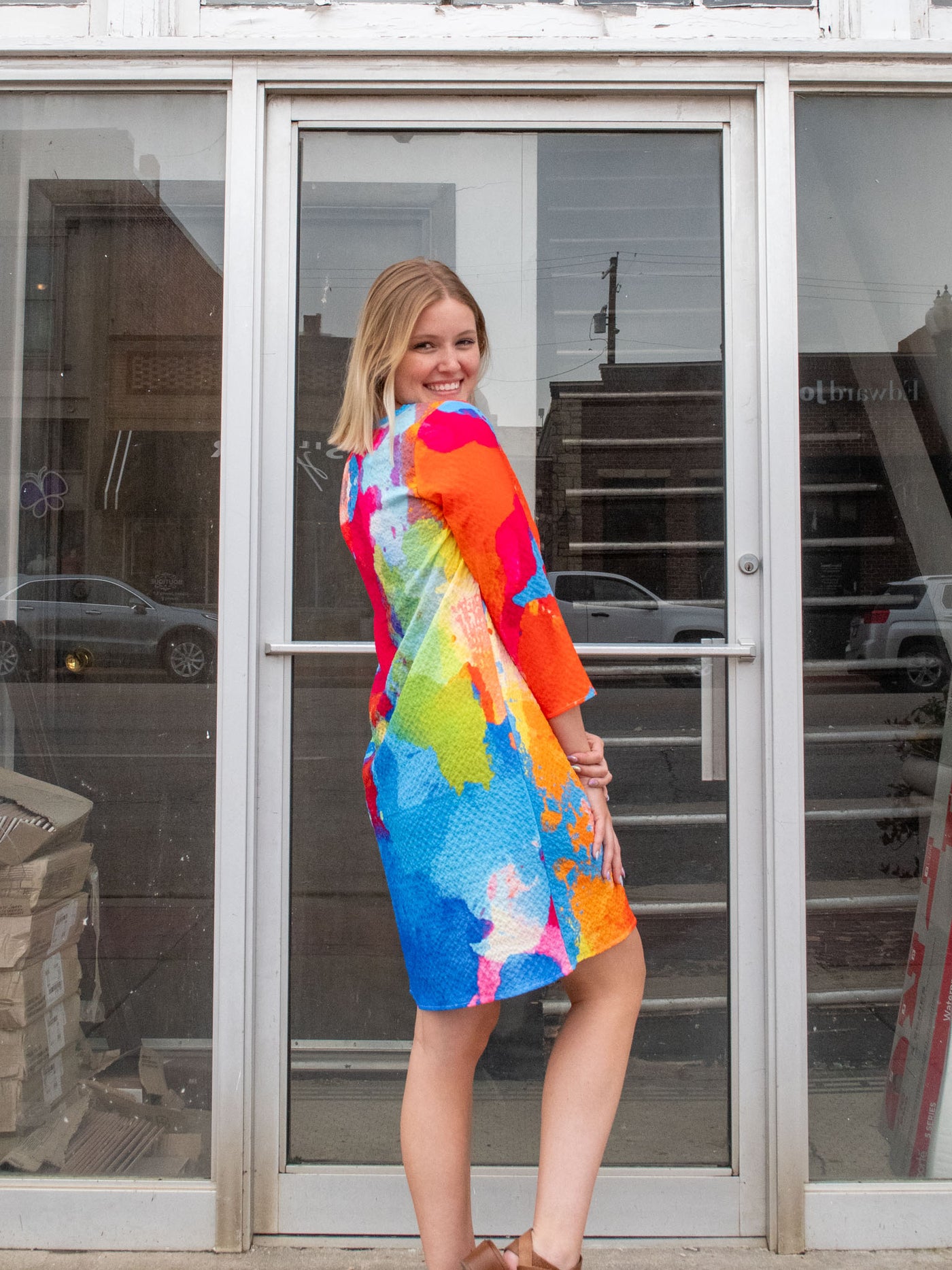 A model wearing a mid thigh length dress that features pink, blue, green, orange, and yellow watercolor details and 3/4 sleeve length. The model paired it with brown platform sandals.