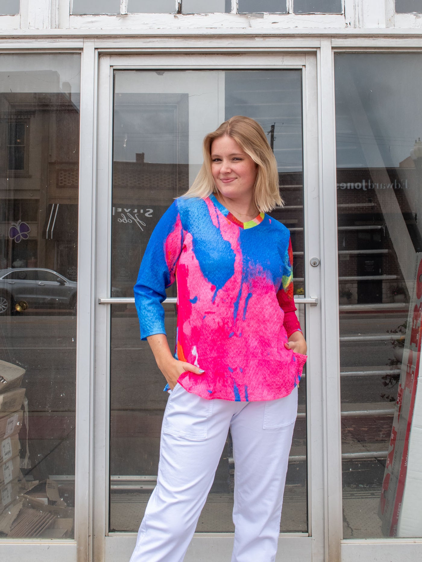 A model wearing a v-neck blouse tee that has a pink, orange, blue, and red pattern and 3/4 sleeve. The model paired it with white drawstring joggers. 