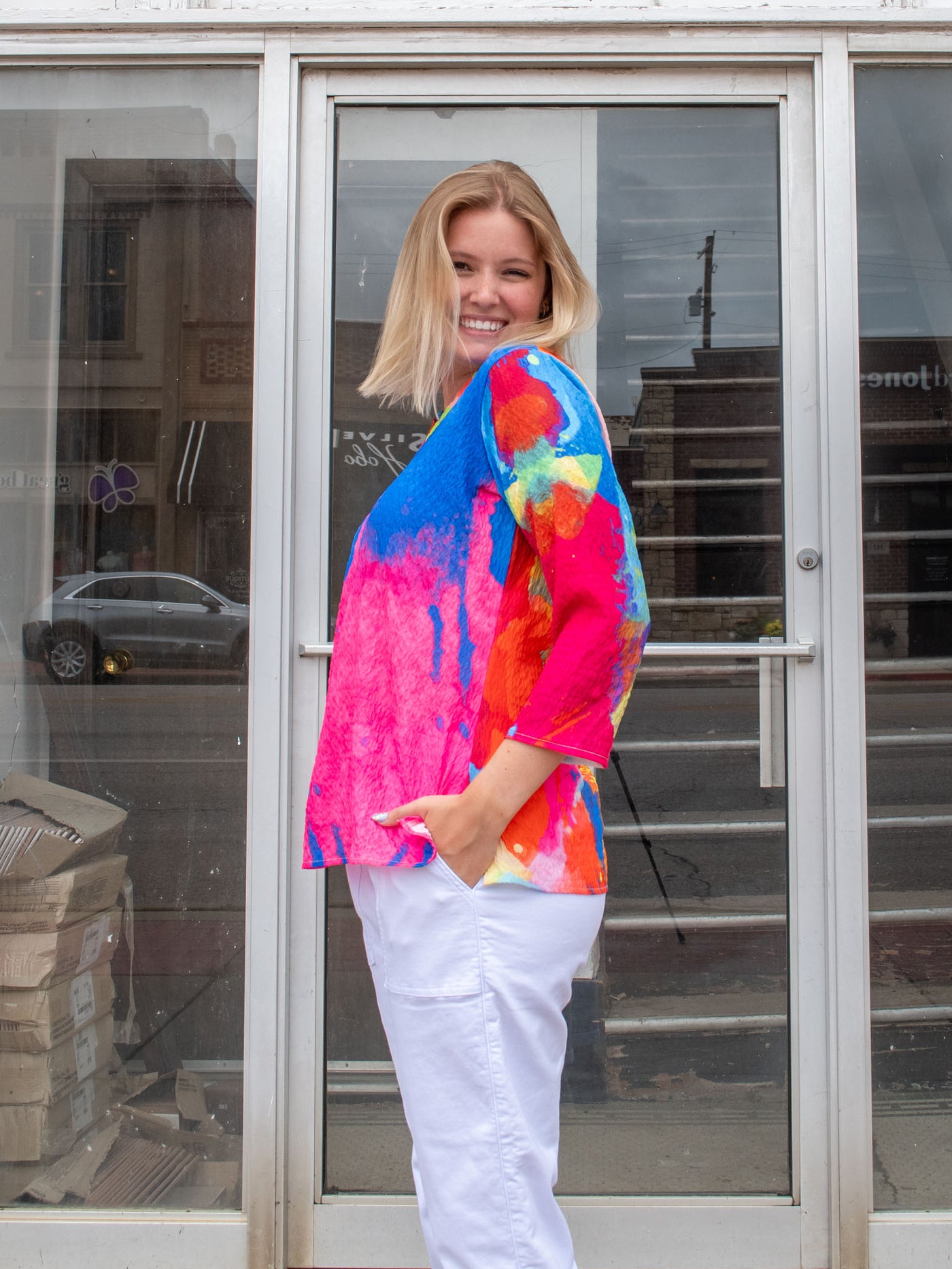 A model wearing a v-neck blouse tee that has a pink, orange, blue, and red pattern and 3/4 sleeve. The model paired it with white drawstring joggers. 