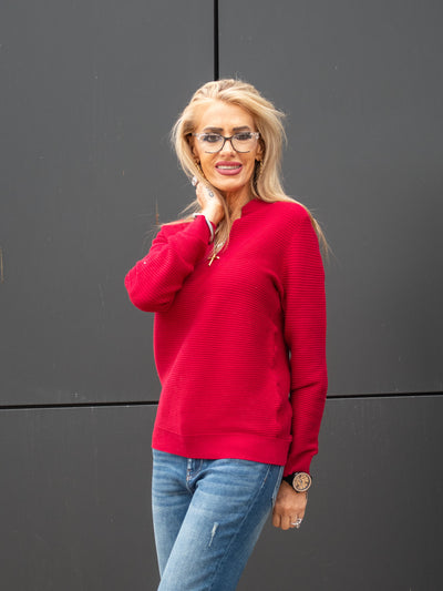A model wearing a red ribbed v-neck, long sleeve top. The model has it paired with a medium wash jean.