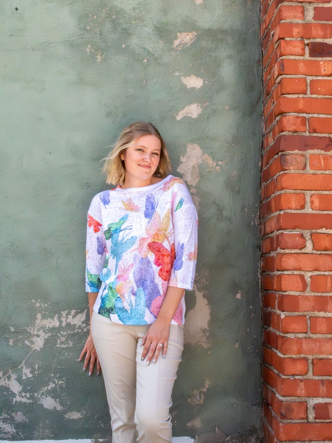 A model wearing a white top with a colorful cascading butterfly pattern. The model has it paired with a pair of white slacks.