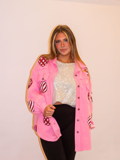 A model wearing a pink button up shacket with sequin Christmas ornaments on the front and sleeves. The model has it paired over a white sequin top and black leggings.