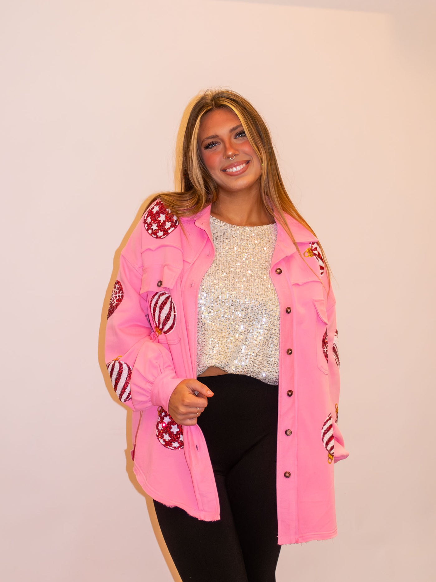 A model wearing a pink button up shacket with sequin Christmas ornaments on the front and sleeves. The model has it paired over a white sequin top and black leggings.