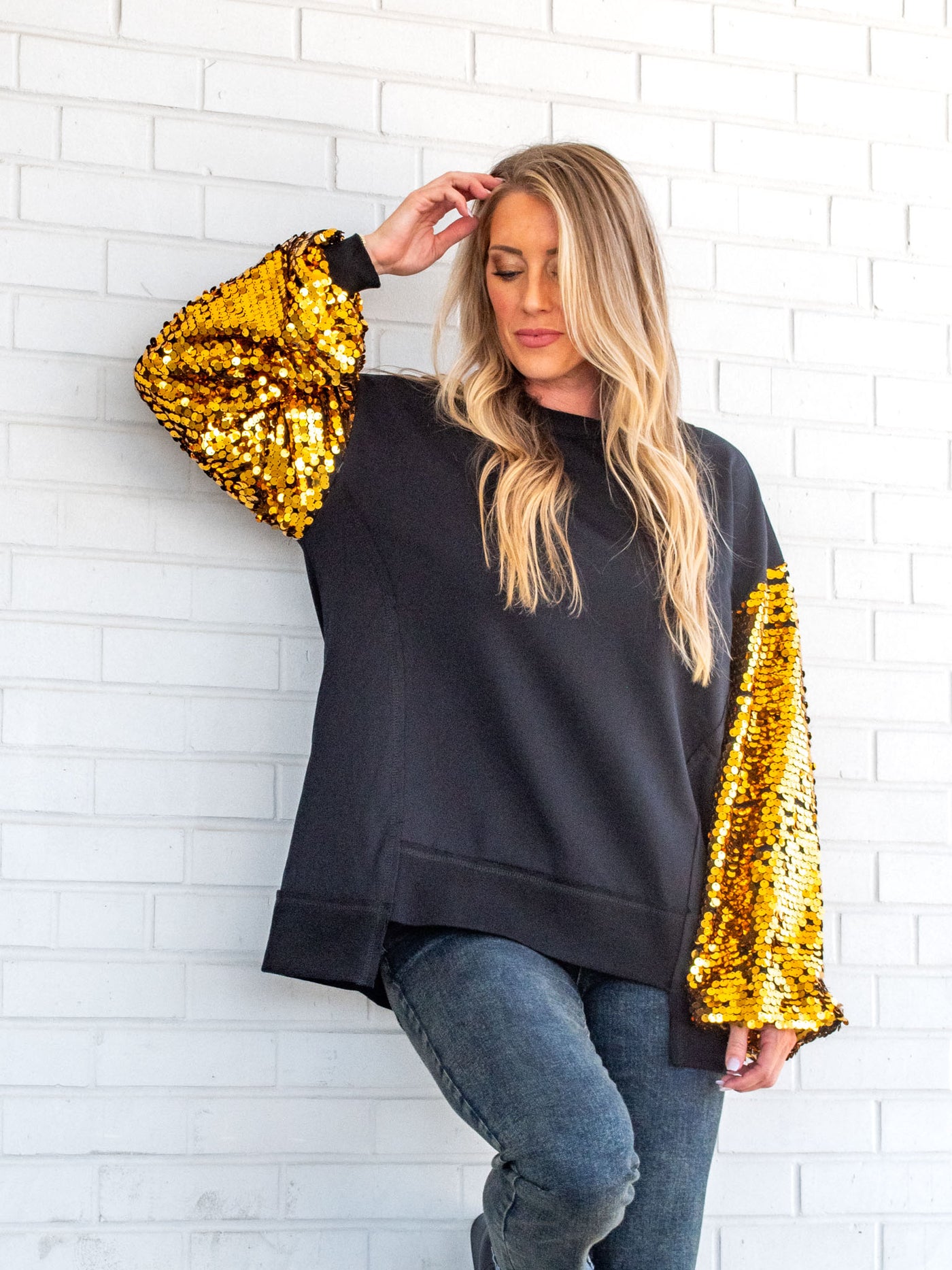A model wearing a black sweatshirt with gold sequin sleeves. The model has it paired with a gray jean.