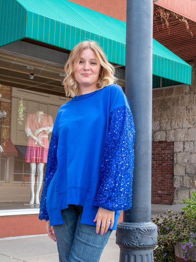 A model wearing a blue oversized sweatshirt with pearl and sparkle details. The model has it paired with a medium wash jean.