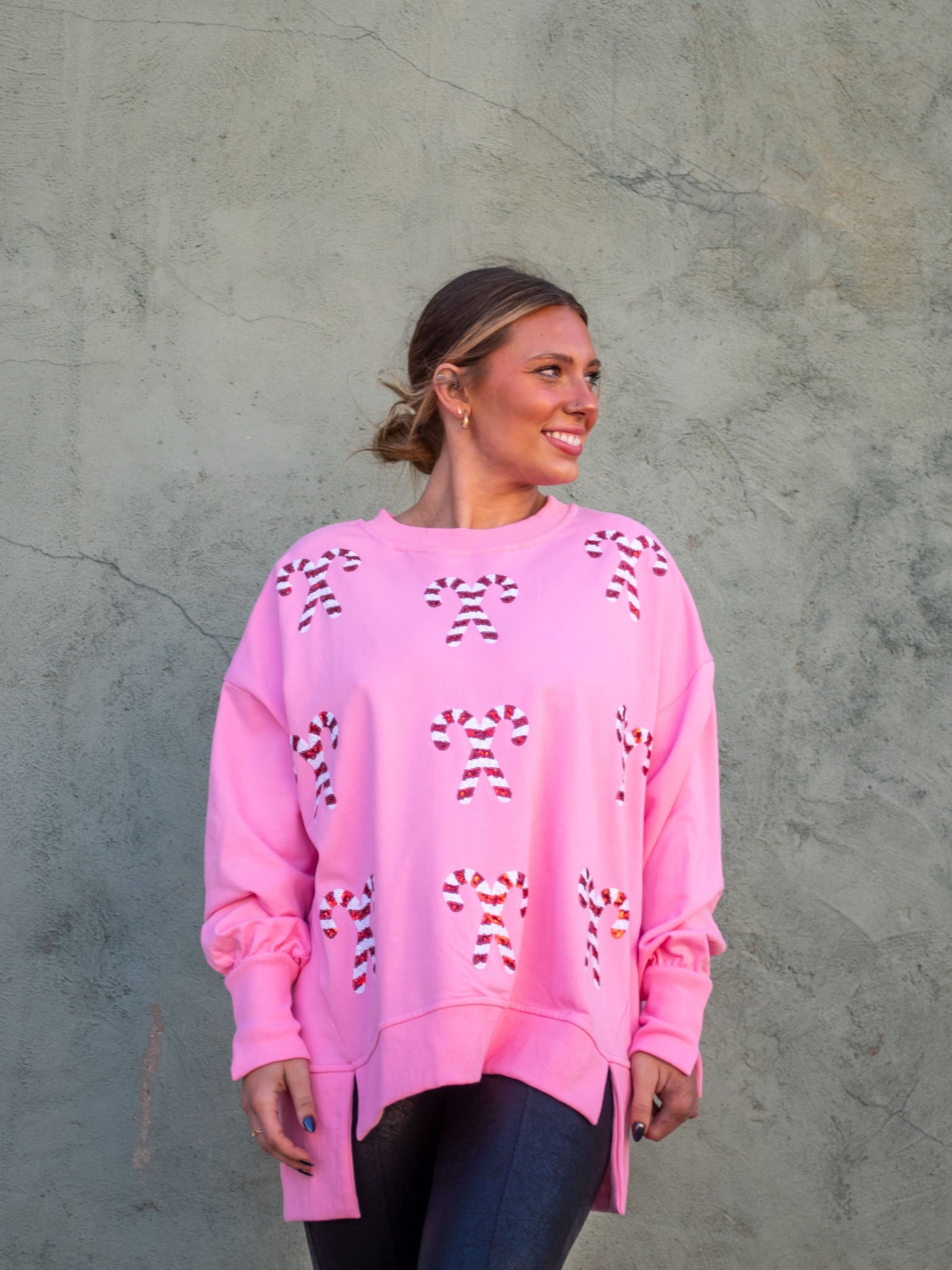 A model wearing a pink oversized sweatshirt with sequin candy cane patches on the front. The model has it paired with a black faux leather legging.