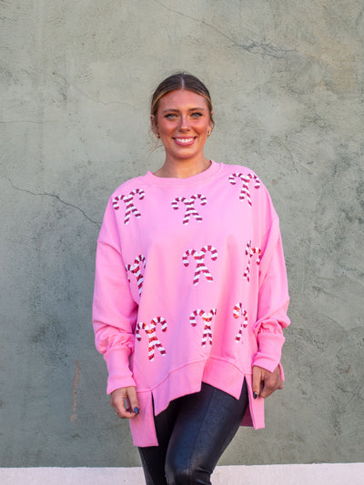 A model wearing a pink oversized sweatshirt with sequin candy cane patches on the front. The model has it paired with a black faux leather legging.