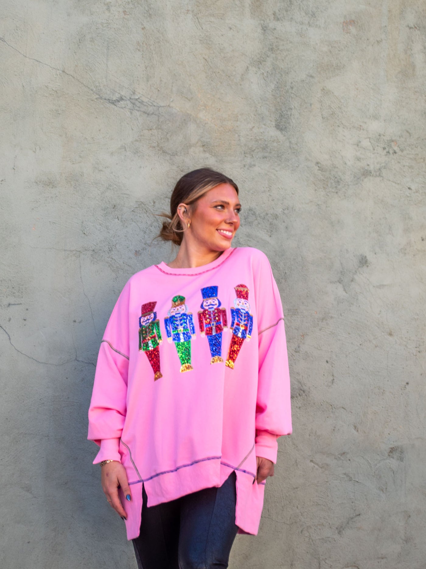 A model wearing an oversized pink sweatshirt with sequin nutcrackers on the front and colorful accent seams. The model has it paired with a faux leather black legging.