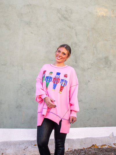 A model wearing an oversized pink sweatshirt with sequin nutcrackers on the front and colorful accent seams. The model has it paired with a faux leather black legging.