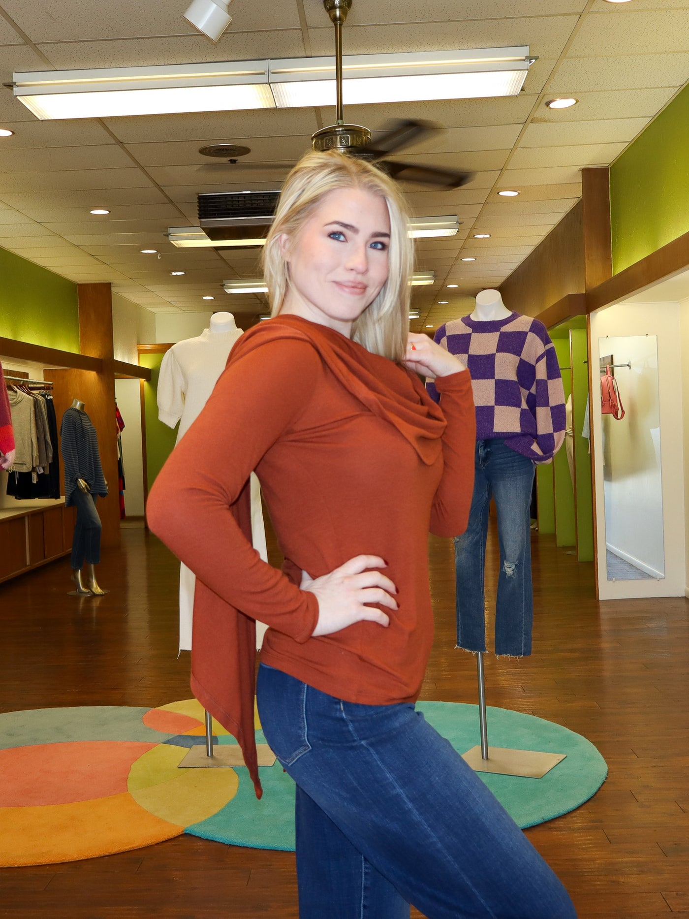 Model is wearing a rust colored reversible longsleeve with a built in shawl that can be reversible to however model wants it to lay. Model has shawl pushed over her shoulders. Long sleeve is paired with blue jeans.