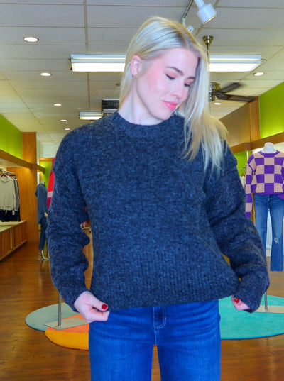 Modeling is wearing a dark heather grey cropped sweater. Paired with medium wash blue jeans. Sweater has small slits on both sides of the sweater at the waist.