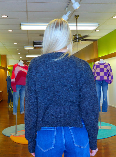 Modeling is wearing a dark heather grey cropped sweater. Paired with medium wash blue jeans. Sweater has small slits on both sides of the sweater at the waist.