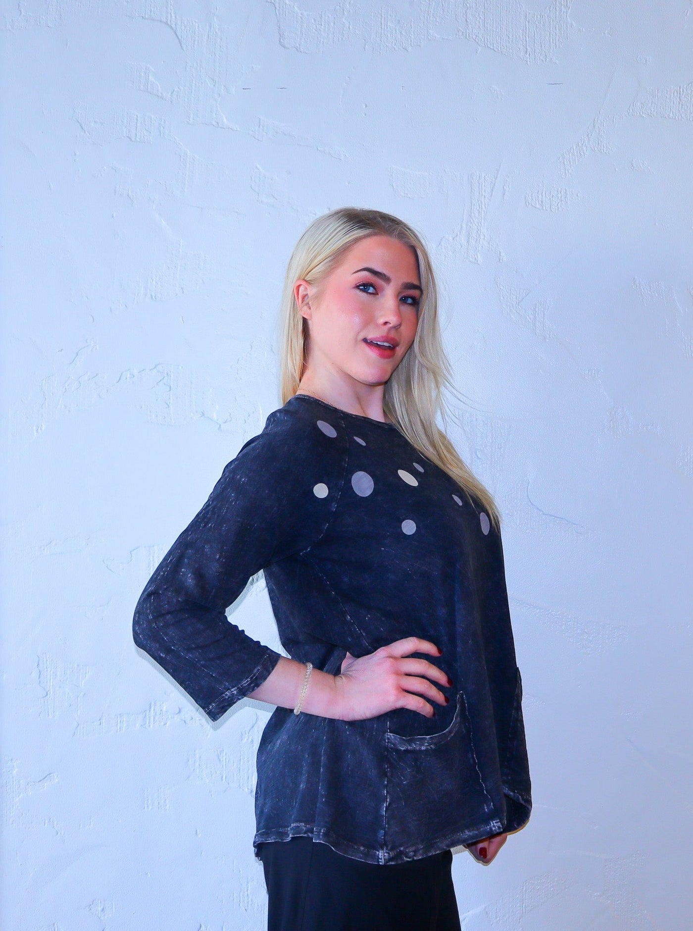 Model is wearing a dark charcoal grey 3/4th sleeve tunic with a distressed print and polka dots at the chest area of garment. Tunic has two pockets on both sides on front of garment. Shirt is paired with black joggers.