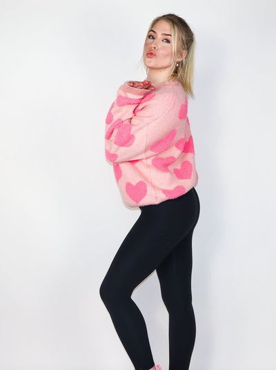 Model is wearing a peach colored knitted sweater with hot pink hearts. Sweater is paired with black leggings.