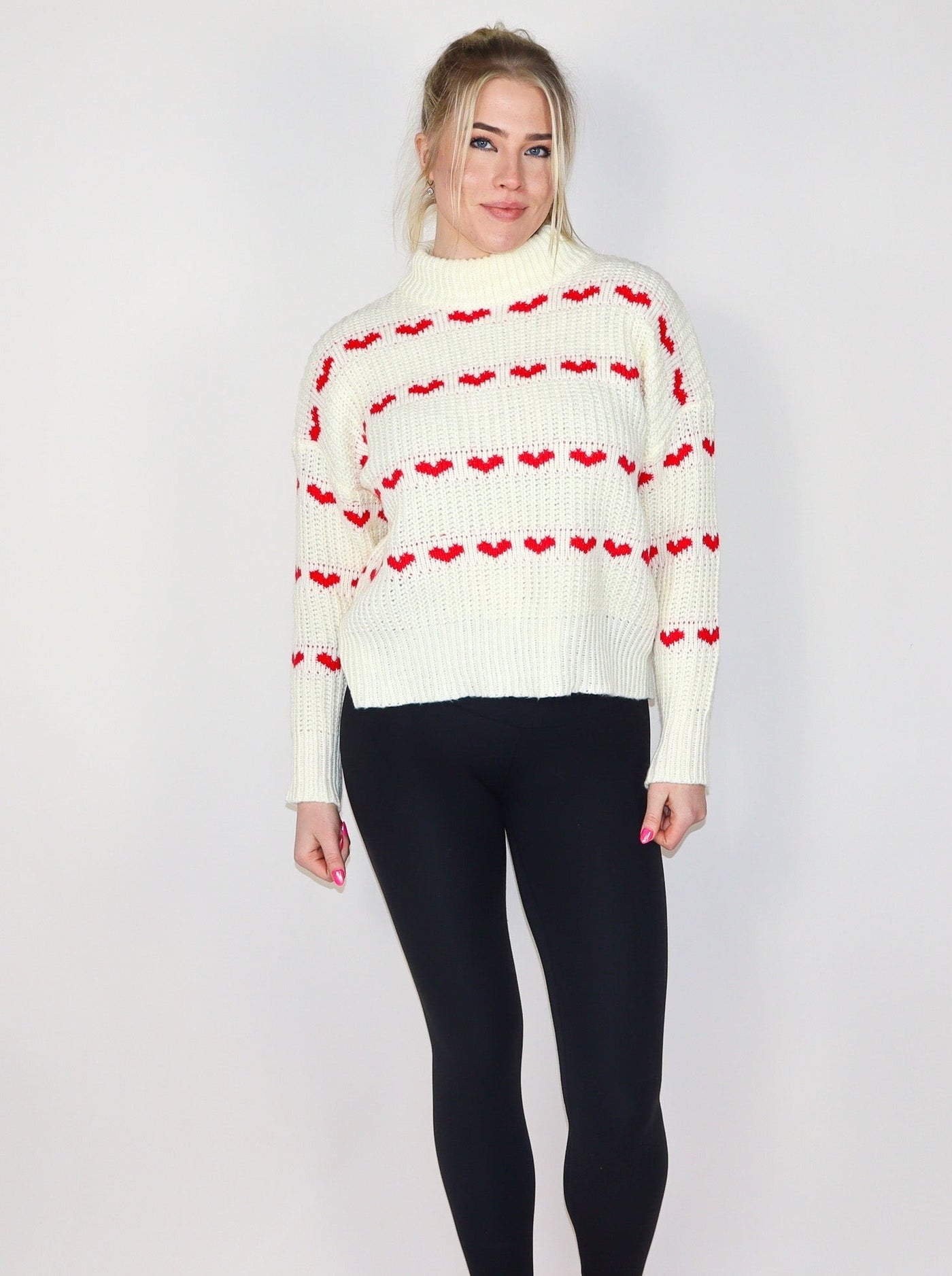 Model is wearing a white knitted turtle neck sweater with horizontal rows of small red hearts. 