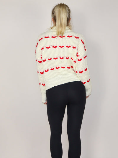 Model is wearing a white knitted turtle neck sweater with horizontal rows of small red hearts.