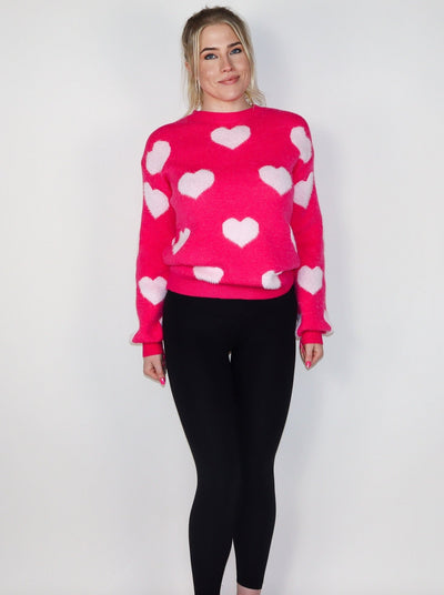 Model is wearing a dark hot pink knitted sweater with light pink hearts. Sweater is paired with black leggings. 