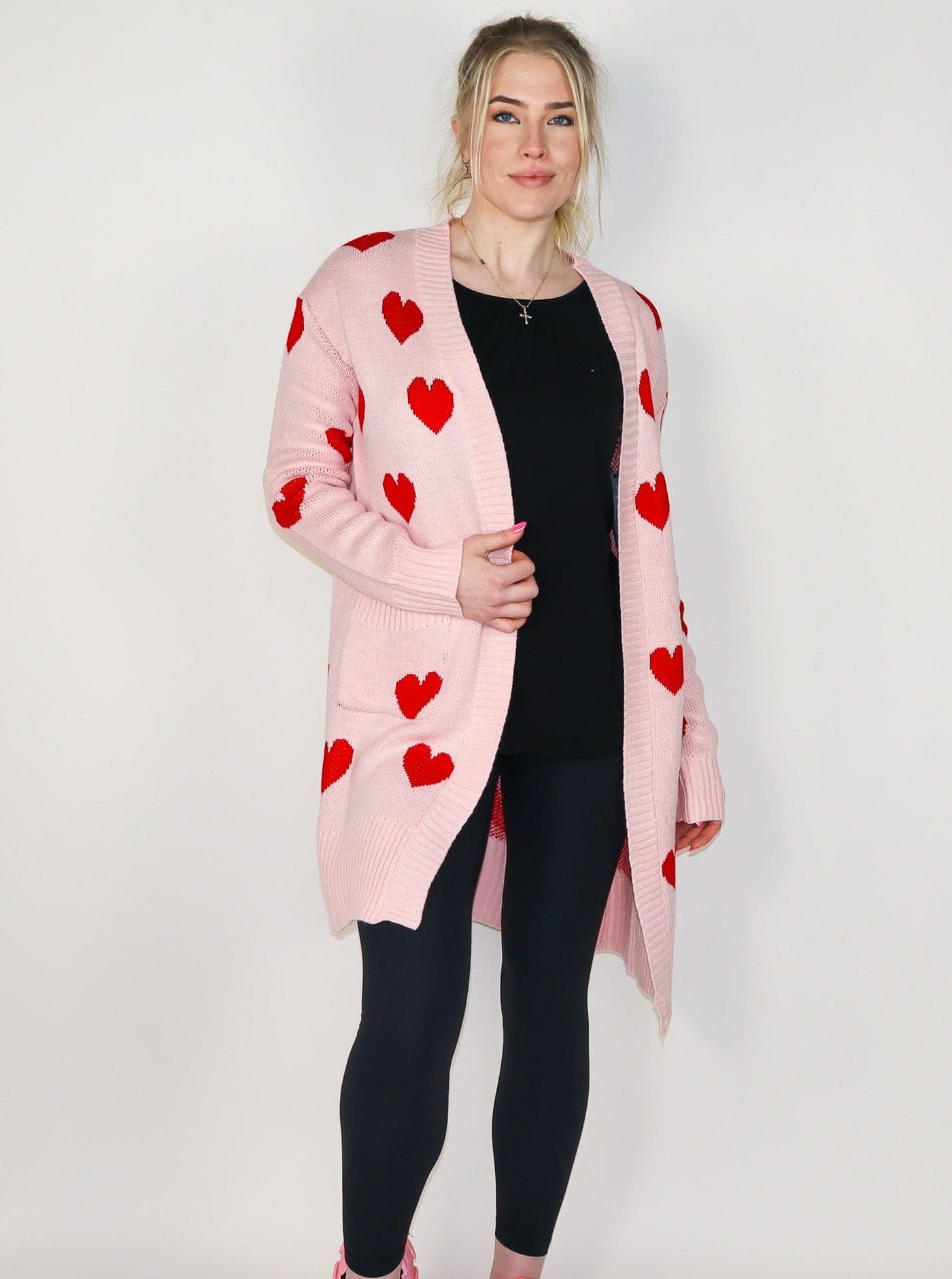 Model is wearing a midi length pink cardigan with red hearts. Cardigan is paired with a black tank top and black leggings. 