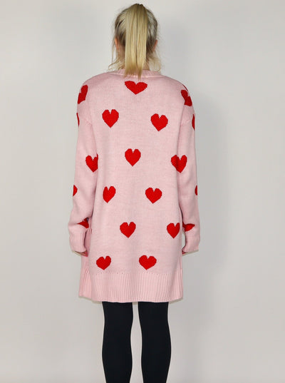 Model is wearing a midi length pink cardigan with red hearts. Cardigan is paired with a black tank top and black leggings.