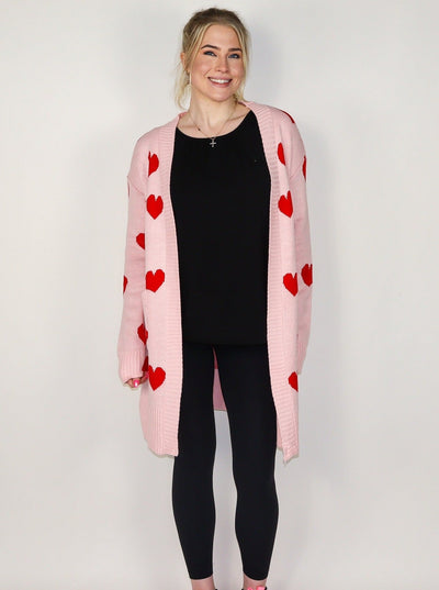 Model is wearing a midi length pink cardigan with red hearts. Cardigan is paired with a black tank top and black leggings.
