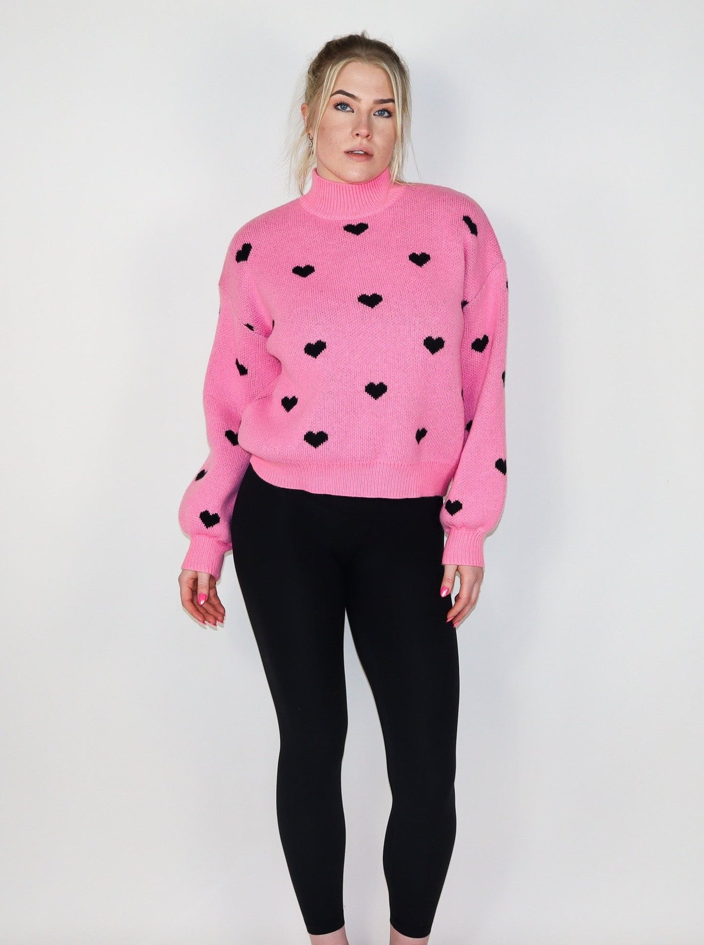 Model is wearing a hot pink turtle neck knitted sweater with black hearts. Sweater is paired with black leggings. 
