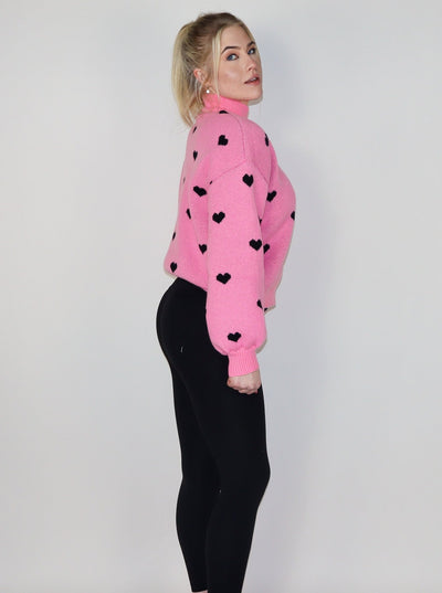 Model is wearing a hot pink turtle neck knitted sweater with black hearts. Sweater is paired with black leggings. 