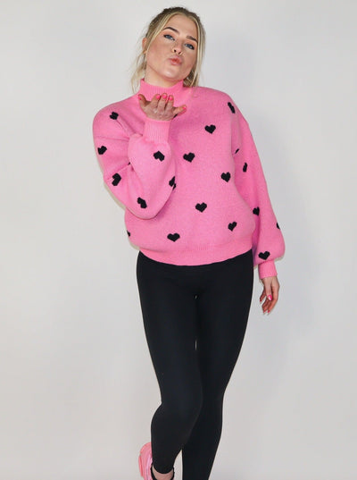 Model is wearing a hot pink turtle neck knitted sweater with black hearts. Sweater is paired with black leggings.