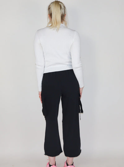 Model is wearing 3/4th length black cargo pants. Pants have 2 sets of pockets, one pair at waist and the other pair on the side, mid thigh. Cargo Pants are paired with a white turtle neck and pink tennis shoes.