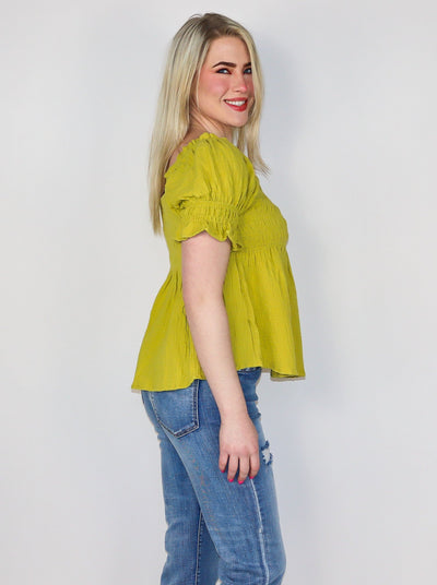 Model is wearing a yellow green blouse with cinching on the bust and sleeves. Boddess is flowey. Top has a square neckline. Top is paired with blue jeans.
