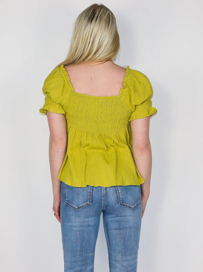 Model is wearing a yellow green blouse with cinching on the bust and sleeves. Boddess is flowey. Top has a square neckline. Top is paired with blue jeans.