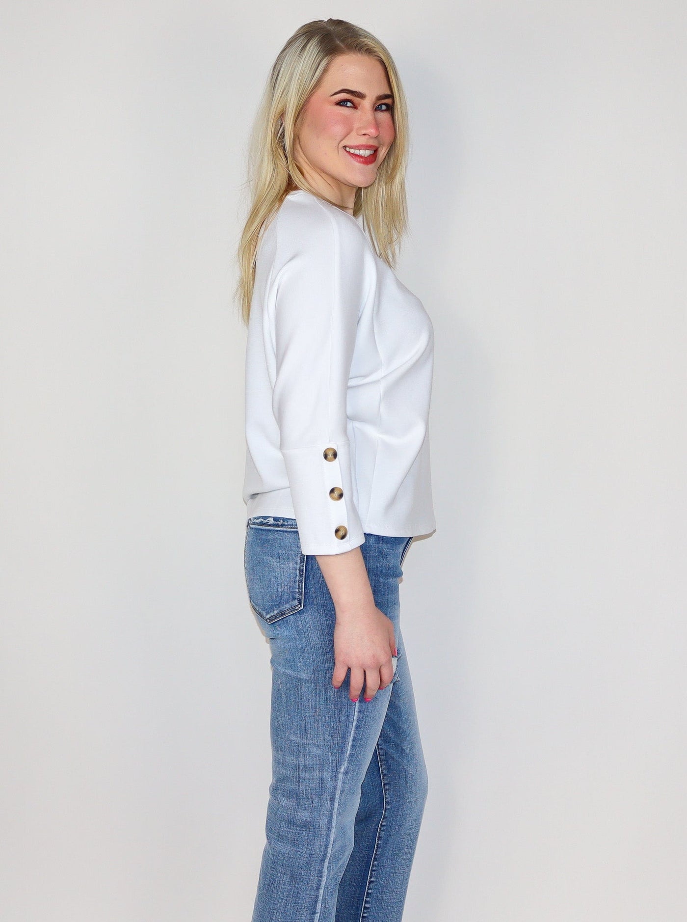 Model is wearing a white 3/4th sleeve top with 3 brown buttons on each sleeve. Top is worn with blue jeans.
