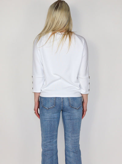 Model is wearing a white 3/4th sleeve top with 3 brown buttons on each sleeve. Top is worn with blue jeans.