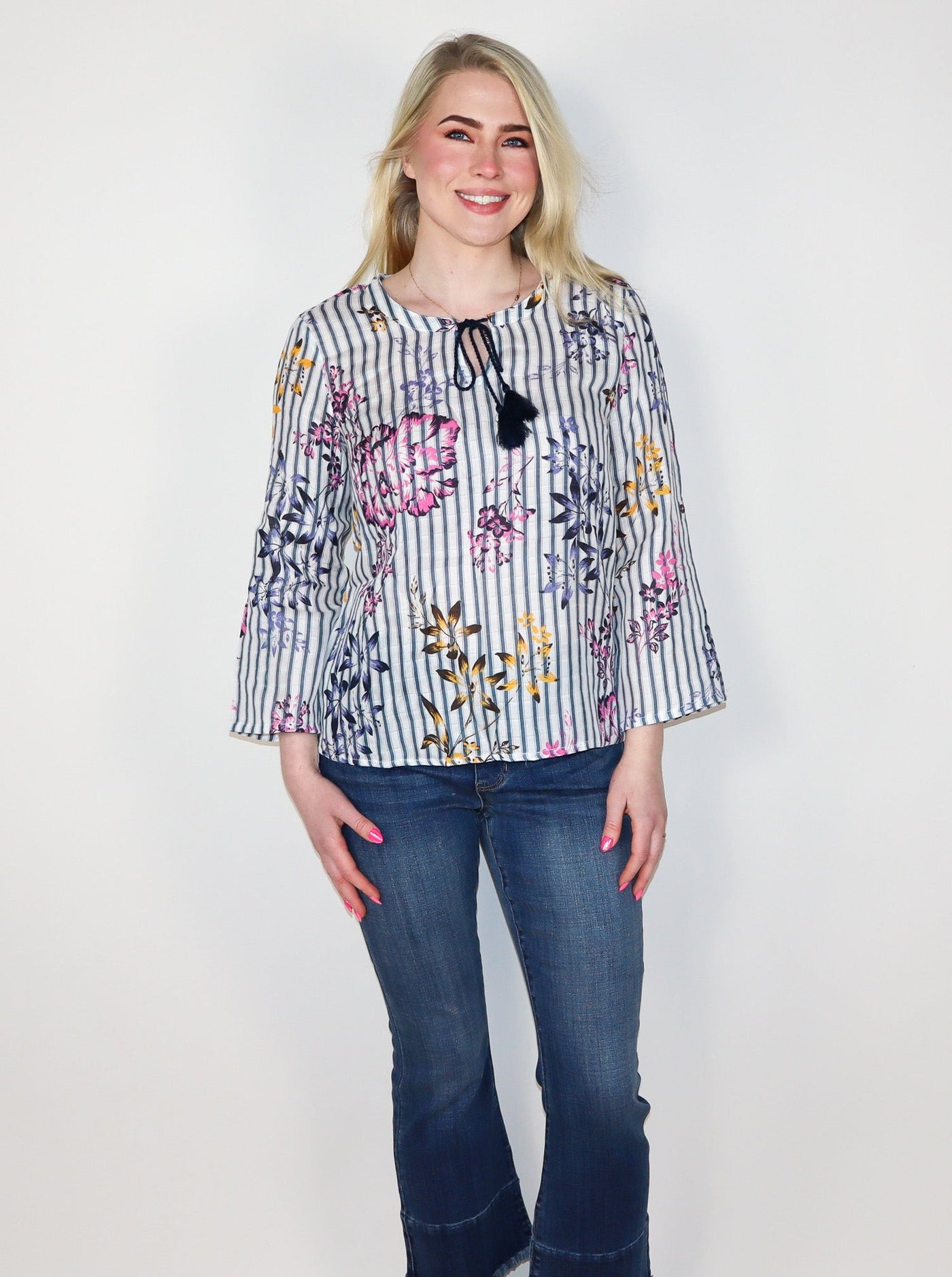 Model is wearing a vertical striped long sleeve blouse with multi color floral detail. The blouse has a string on the neckline for a bowtie detail. Blouse is paired with blue jeans. 
