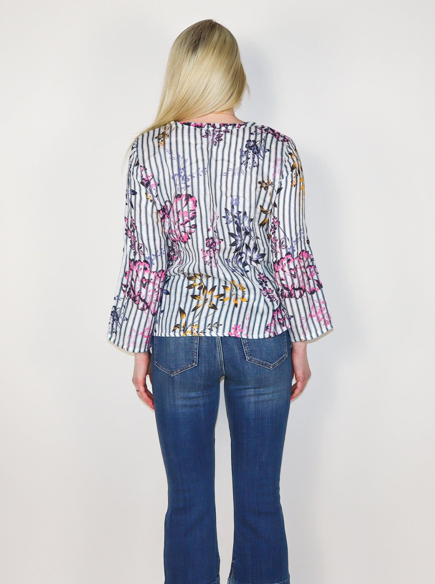 Model is wearing a vertical striped long sleeve blouse with multi color floral detail. The blouse has a string on the neckline for a bowtie detail. Blouse is paired with blue jeans.