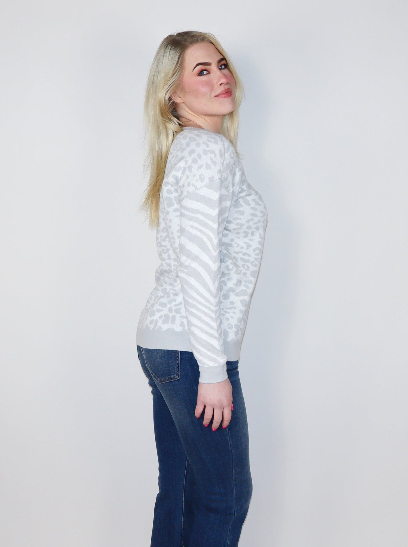 Model is wearing a white and light grey cheetah and zebra print pullover. Pullover is paired with blue jeans.