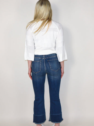 Model is wearing a dark wash 3/4th length jean with frayed detail on hemming and stitching horizontal across the bottom of shin. Jeans are paired with a white long sleeve blouse. 