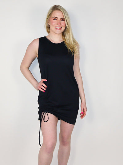 Model is wearing a fitted black sleeveless shift dress with shirring on the right thigh.  Dress is worn with white tennis shoes. 