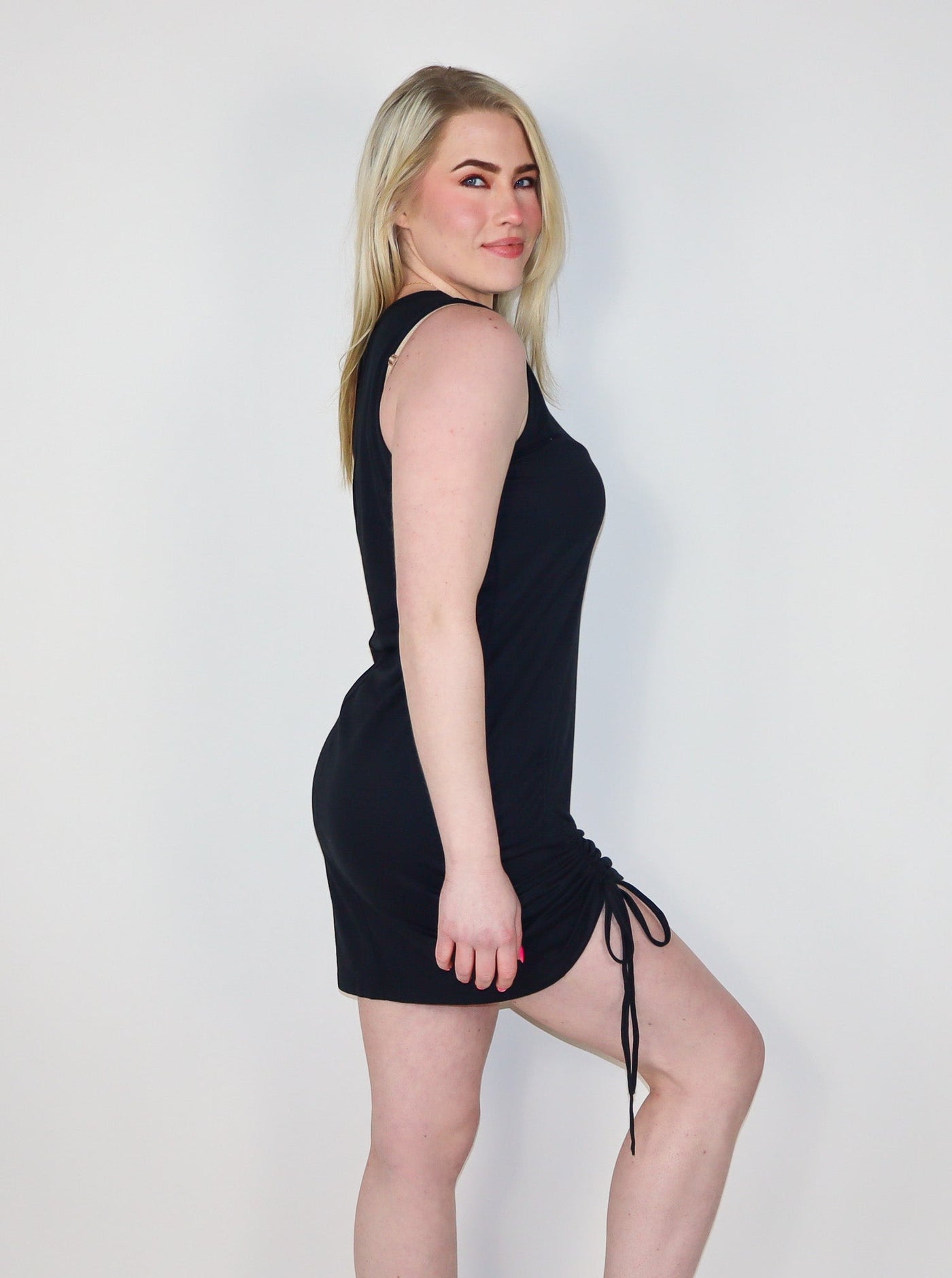 Model is wearing a fitted black sleeveless shift dress with shirring on the right thigh. Dress is worn with white tennis shoes.