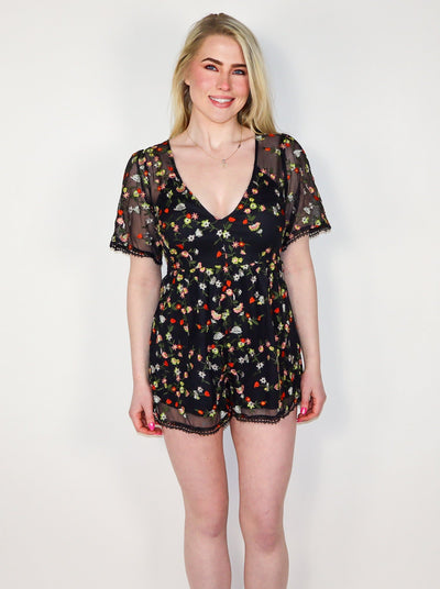 Model is wearing a sheer fitted romper with flutter sleeves and floral detail. Neckline is a deep v-neck. 