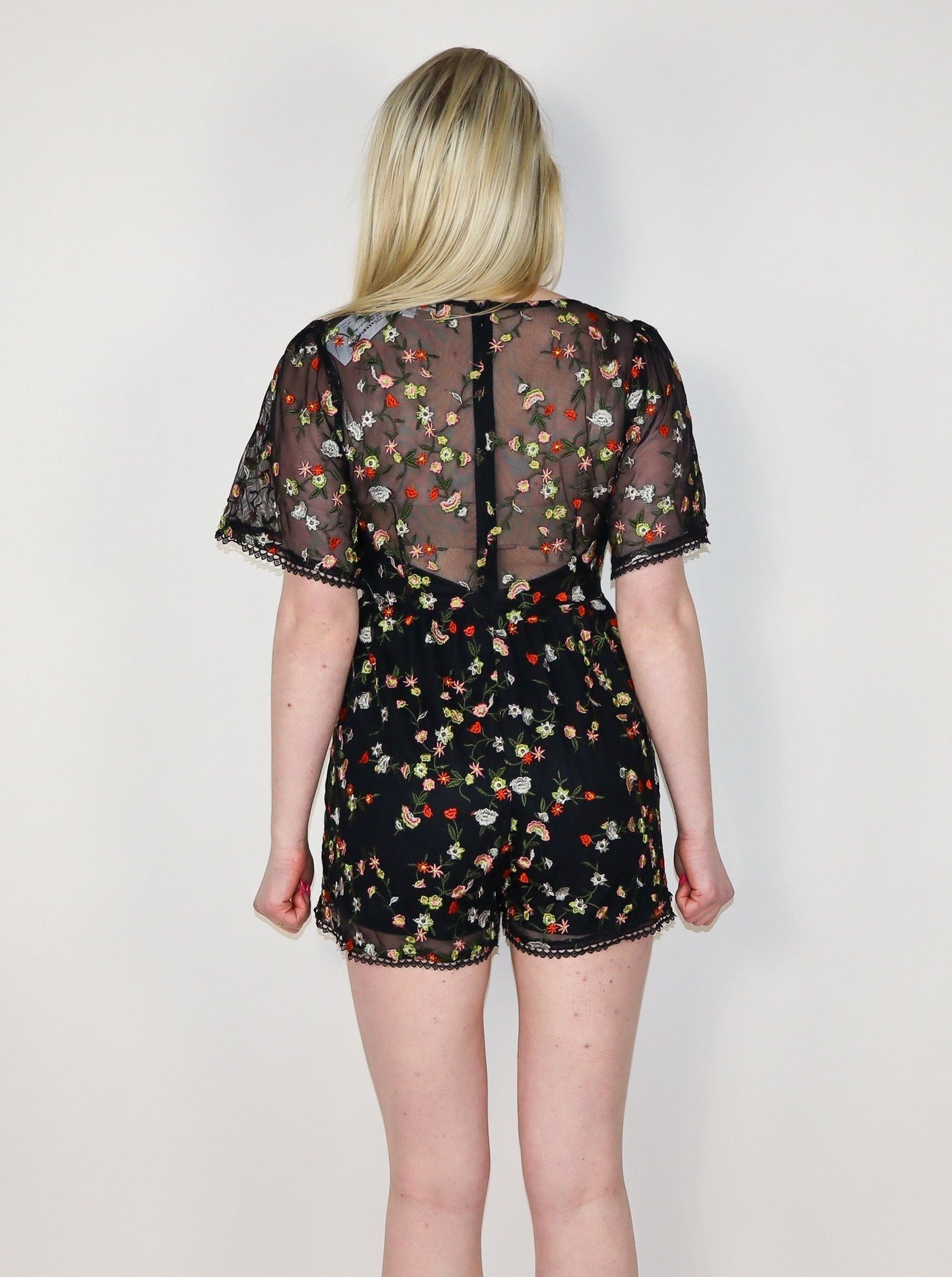 Model is wearing a sheer fitted romper with flutter sleeves and floral detail. Neckline is a deep v-neck.