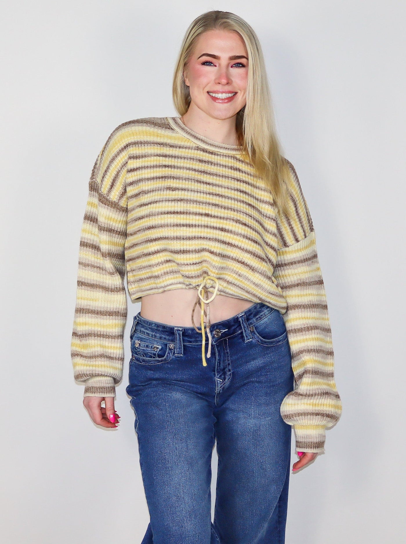 Model is wearing a yellow and light brown stripped cropped sweater with a drawstring at waist worn with blue jeans. 