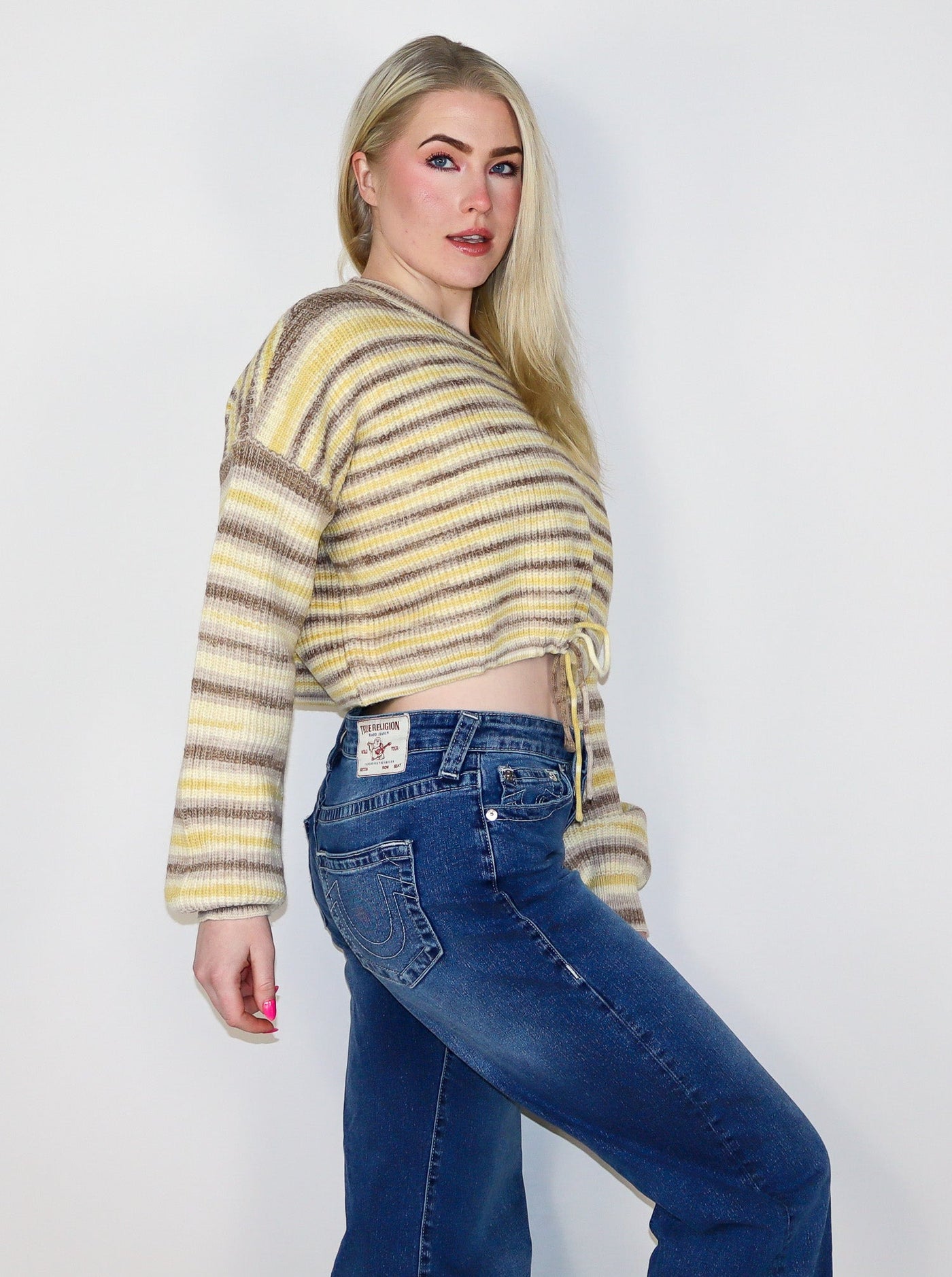 Model is wearing a yellow and light brown stripped cropped sweater with a drawstring at waist worn with blue jeans. 