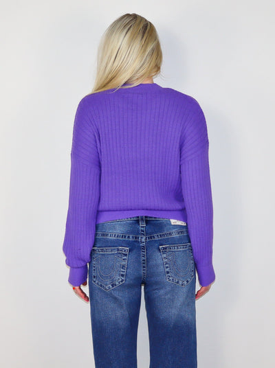Model is wearing a ribbed purple sweater with blue jeans. 