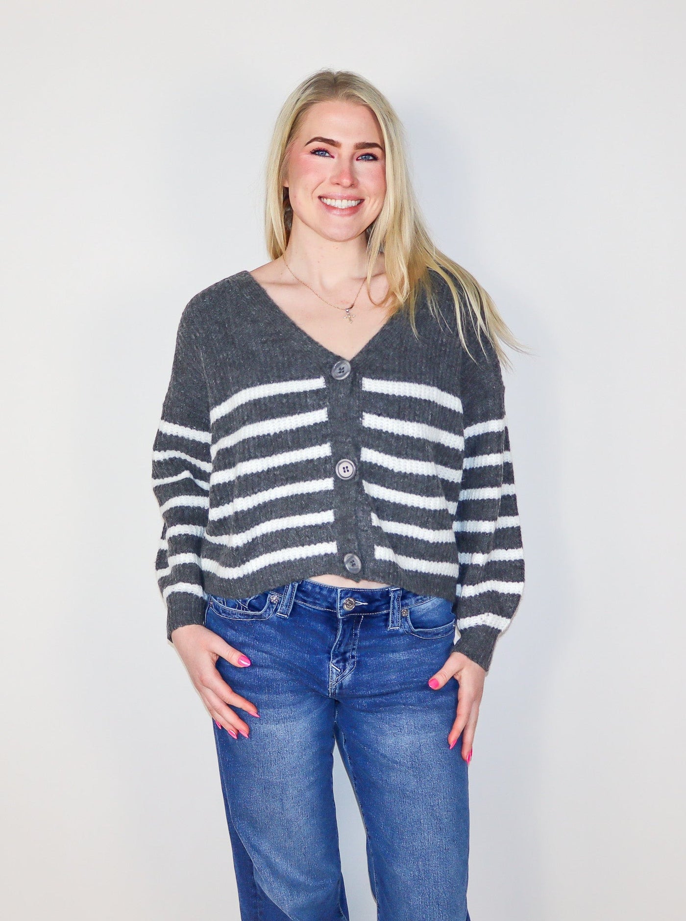 Model is wearing a grey and white horizontal striped cropped button up sweater with a v-neck line. Sweater is paired with blue jeans. 