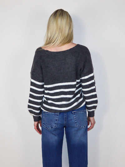 Model is wearing a grey and white horizontal striped cropped button up sweater with a v-neck line. Sweater is paired with blue jeans. 