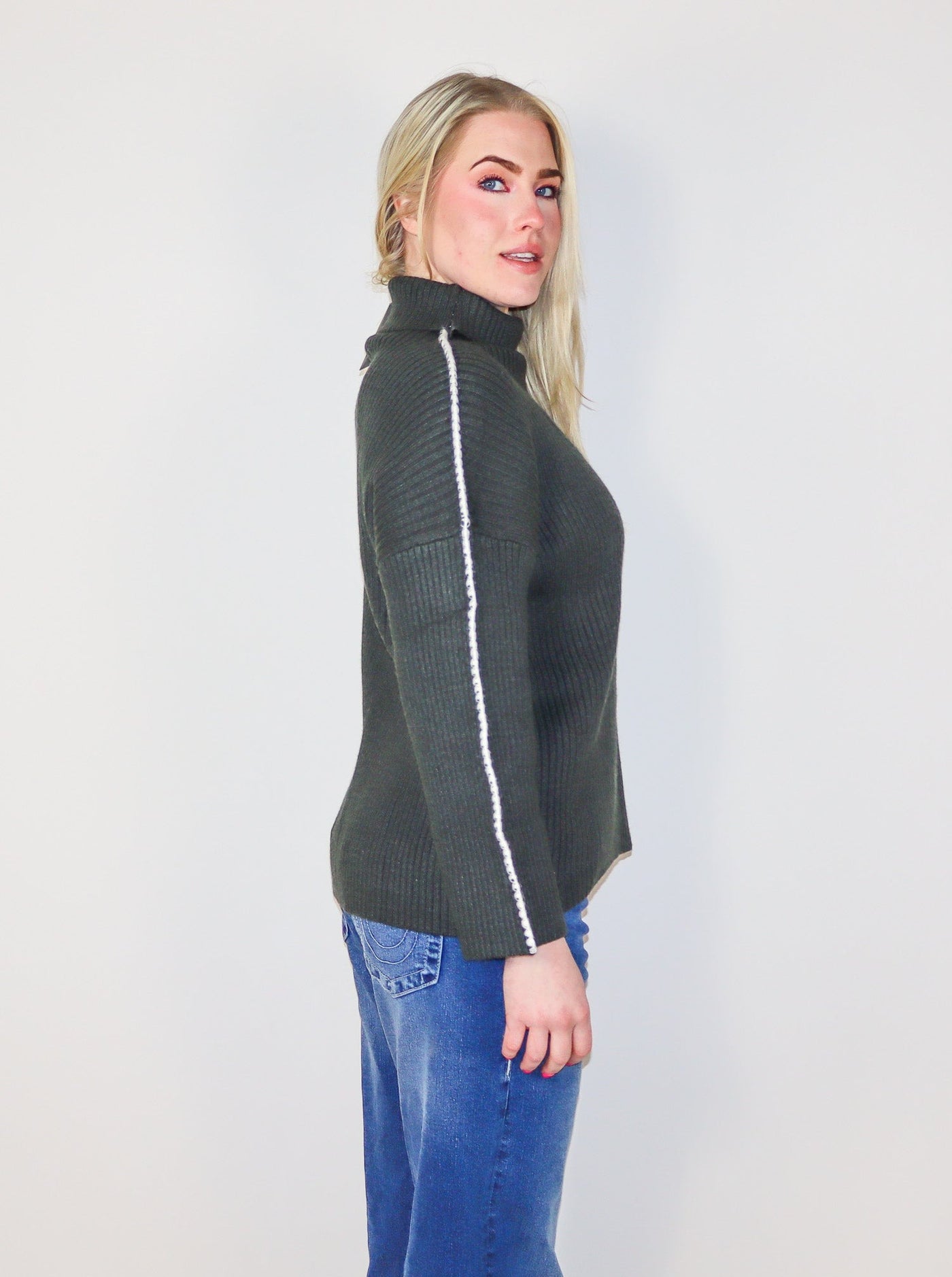 Model is wearing a dark charcoal colored turtle neck sweater with white stitching on the sleeves. 