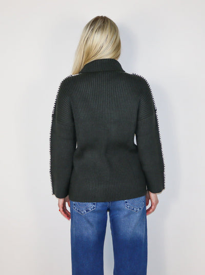Model is wearing a dark charcoal colored turtle neck sweater with white stitching on the sleeves. 